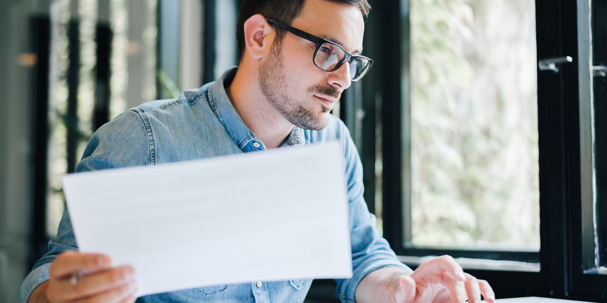 Man with glasses typing on a laptop with a paper in his hand