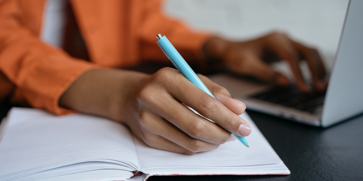 A woman simultaneously taking notes while working on her laptop at a desk.