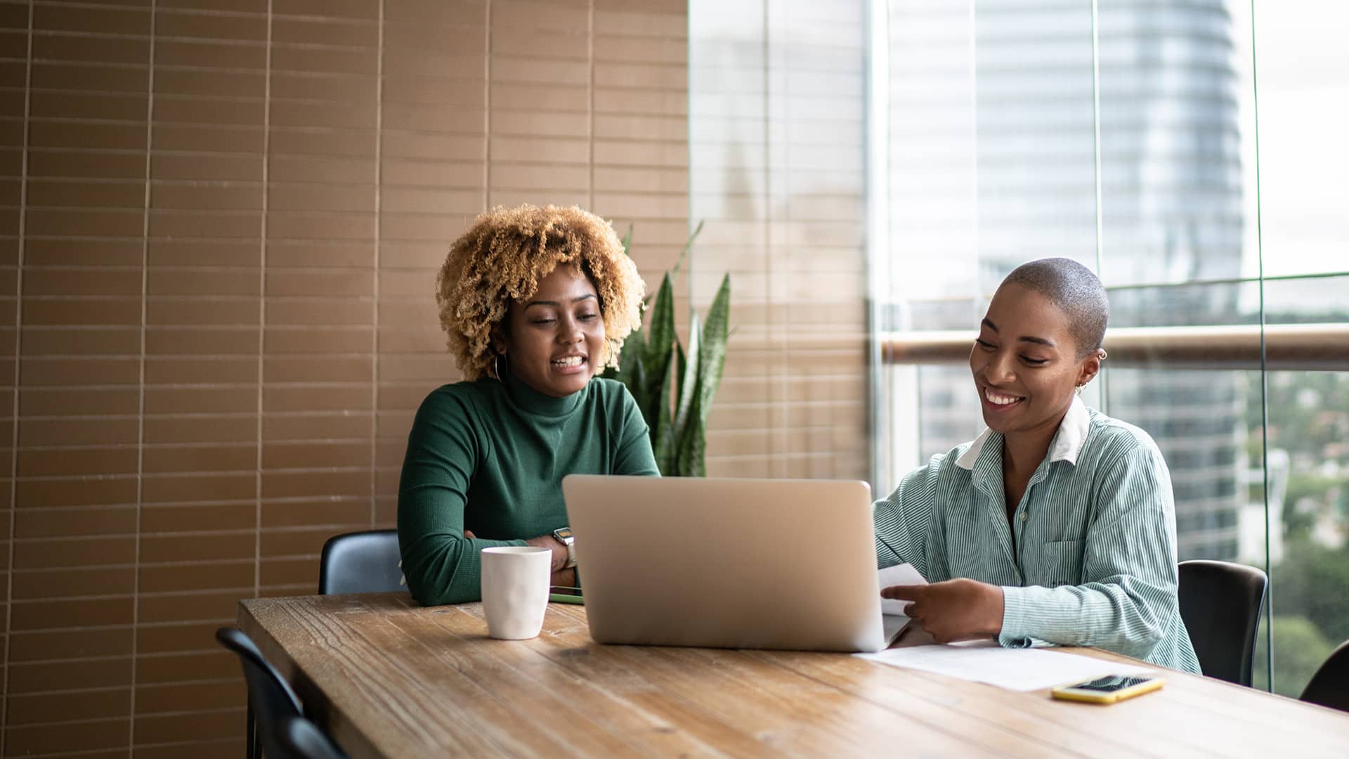 Two women smiling while working on a laptop together in a modern office.